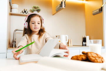 Little girl, pupil wearing headphones, listening to lesson, doing homework, sitting in kitchen