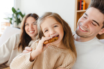 Beautiful smiling girl, mom, dad and cute little girl eating cookies, looking at camera in cozy home - obrazy, fototapety, plakaty