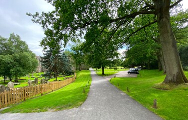 Pathway in Bolton Abbey, with an old graveyard, grassland, trees, and distant Abbey buildings in, Bolton Abbey, UK