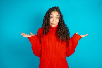 Clueless Beautiful teen girl wearing knitted red sweater over blue background shrugs shoulders with hesitation, faces doubtful situation, spreads palms, Hard decision - obrazy, fototapety, plakaty