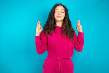 Beautiful young girl wearing pink tracksuit on blue background has big hope, crosses fingers,...