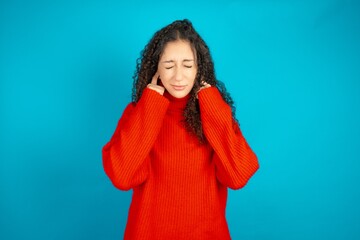 beautiful teen girl wearing red knitted sweater covering ears with fingers with annoyed expression...