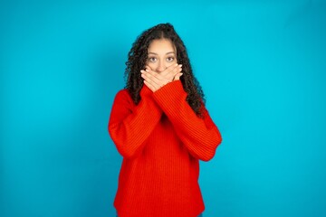 Beautiful teen girl wearing knitted red sweater over blue background shocked covering mouth with...
