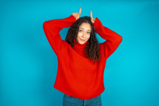 Funny Beautiful teen girl wearing knitted red sweater over blue background shows horns, fingers on head gesture, posing silly and cute