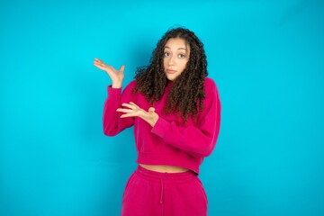 Beautiful young girl wearing pink tracksuit on blue background pointing aside with both hands...