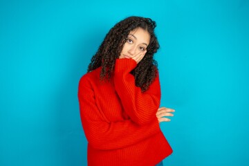 Very bored Beautiful teen girl wearing knitted red sweater over blue background holding hand on...