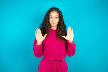 Serious Beautiful young girl wearing pink tracksuit on blue background pulls palms towards camera, makes stop gesture, asks to control your emotions and not be nervous