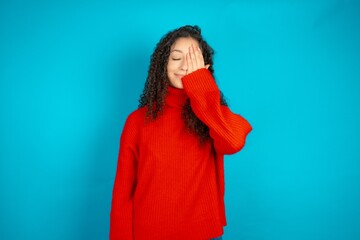 Beautiful teen girl wearing knitted red sweater over blue background covering one eye with her...
