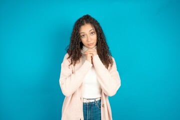 Positive beautiful teen girl wearing brown cardigan smiles happily, glad to receive pleasant news from interlocutor, keeps hands together. People emotions concept.