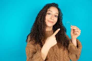 beautiful teen girl wearing brown knitted sweater holding an invisible aligner and pointing at it. Dental healthcare and confidence concept.
