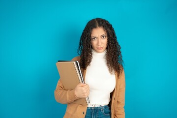 Portrait of dissatisfied beautiful teen woman wearing beige knitted jacket smirks face, purses lips and looks with annoyance at camera, discontent hearing something unpleasant - obrazy, fototapety, plakaty