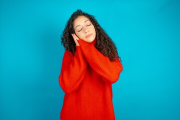 Beautiful teen girl wearing knitted red sweater over blue background sleeping tired dreaming and...