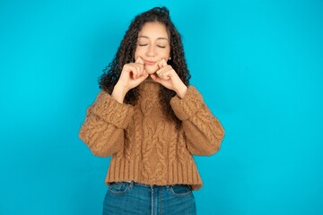 Pleased beautiful teen girl wearing brown knitted sweater  with closed eyes keeps hands near cheeks and smiles tenderly imagines something very pleasant