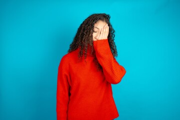 beautiful teen girl wearing red knitted sweater with sad expression covering face with hands while...