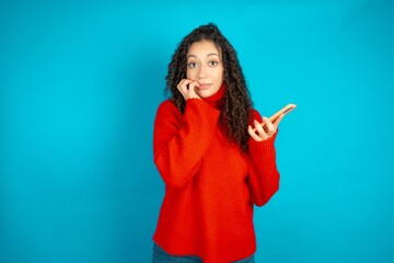 Afraid funny beautiful teen girl wearing red knitted sweater holding telephone and bitting nails - obrazy, fototapety, plakaty