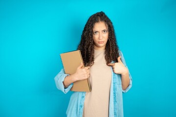 Fototapeta premium Embarrassed beautiful arab student carries notebooks indicates at herself with puzzled expression, being shocked to be chosen to participate in competition, hesitates about something