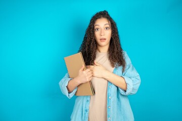 Fototapeta premium Scared beautiful arab student carries notebooks looks with frightened expression, keeps hands on chest, being puzzled to notice something strange, People, hush reaction and emotions.