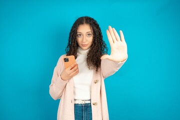 Beautibul teen girl wearing casual clothes  using and texting with smartphone with open hand doing stop sign with serious and confident expression, defense gesture