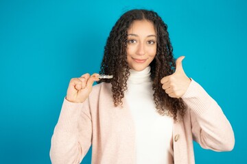 Beautibul teen girl wearing casual clothes holding an invisible braces aligner and rising thumb up,...