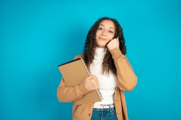 Fototapeta premium Happy beautiful arab student carries notebooks keeps fists on cheeks smiles broadly and has positive expression being in good mood