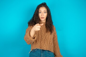Shocked Beautiful teen girl wearing knitted sweater over blue background points at you with stunned...