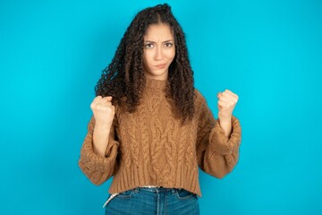 Irritated Beautiful teen girl wearing knitted sweater over blue background blows cheeks with anger and raises clenched fists expresses rage and aggressive emotions. Furious model - Powered by Adobe