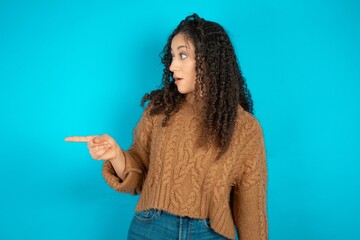 Stunned Beautiful teen girl wearing knitted sweater over blue background with greatly surprised...