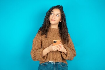 Beautiful teen girl wearing knitted sweater over blue background holds mobile phone uses high speed...