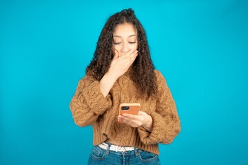 Beautiful teen girl wearing knitted sweater over blue background being deeply surprised, stares at...