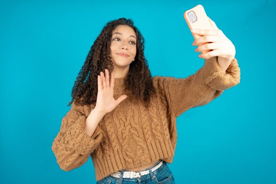 Beautiful teen girl wearing knitted sweater over blue background holds modern mobile phone and makes video call waves palm in hello gesture. People modern technology concept