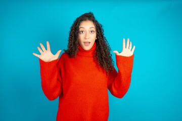Delighted positive beautiful teen girl wearing red knitted sweater opens mouth  and arms palms up...