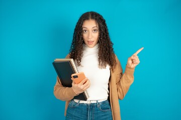 Astonished beautiful caucasian teen girl wearing brown sweater holding her telephone and pointing with finger aside at empty copy space