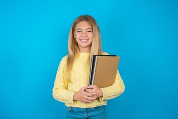 Coquettish Beautiful kid girl wearing yellow sweater holding notebook smiling happily, blinking at camera in a playful manner, flirting with you.