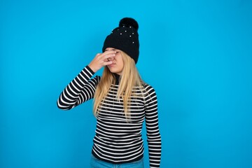 Teen caucasian girl wearing striped sweater and woolly hat smelling something stinky and disgusting, intolerable smell, holding breath with fingers on nose. Bad smell - obrazy, fototapety, plakaty