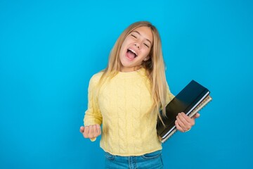 Beautiful kid girl wearing yellow sweater holding notebook celebrating surprised and amazed for success with arms raised and eyes closed. Winner concept.