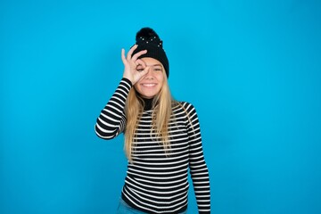 Teen caucasian girl wearing striped sweater and woolly hat doing ok gesture with hand smiling, eye looking through fingers with happy face.