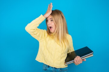 Beautiful kid girl wearing yellow sweater holding notebook surprised with hand on head for mistake,...