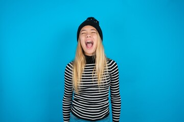 Teen caucasian girl wearing striped sweater and woolly hat angry and mad screaming frustrated and furious, shouting with anger. Rage and aggressive concept.