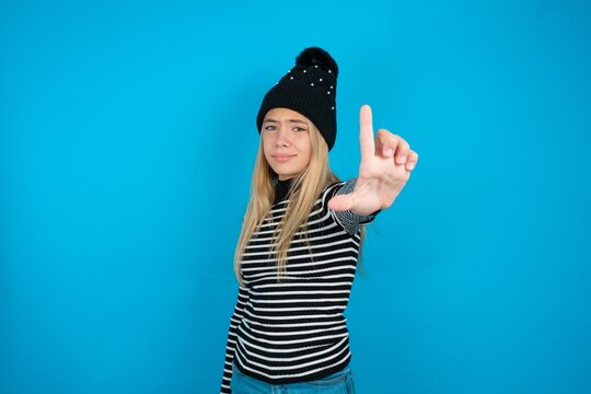 Teen caucasian girl wearing striped sweater and woolly hat making fun of people with fingers on forehead doing loser gesture mocking and insulting.