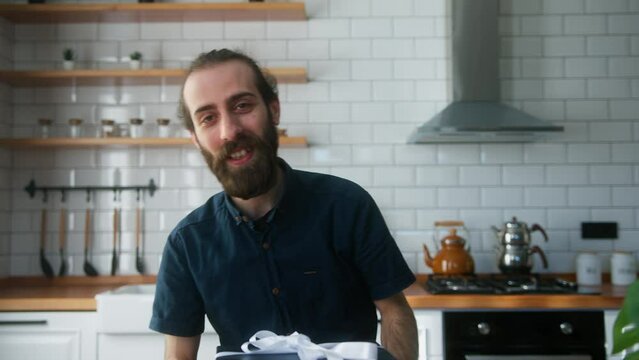 Bearded young vlogger man sitting in kitchen, catch the gift box in the air, advertising product , talking to camera, recording video
