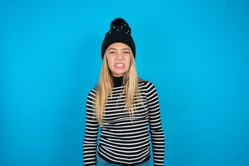 Teen caucasian girl wearing striped sweater and woolly hat keeps teeth clenched, frowns face in...