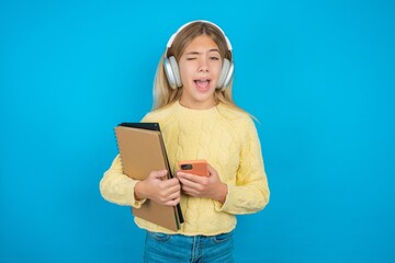 Excited Beautiful kid girl wearing yellow sweater holding notebook winking and eye hold smart phone...