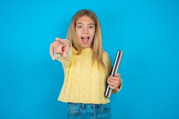 Excited positive beautiful caucasian teen girl wearing yellow sweater points index finger directly at you, sees something very funny. Wow, amazing