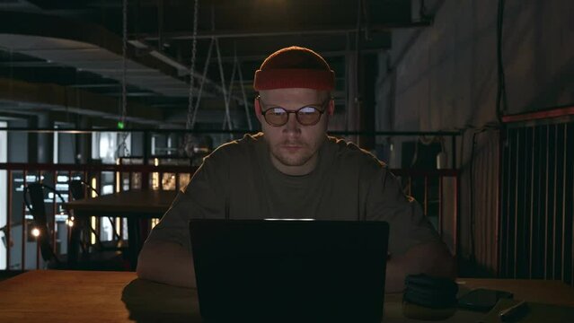 Front view of a 30 year old man with round glasses, who is working on a laptop in a dark cafe room with a calm face. Freelancer in the dark. High quality 4k footage