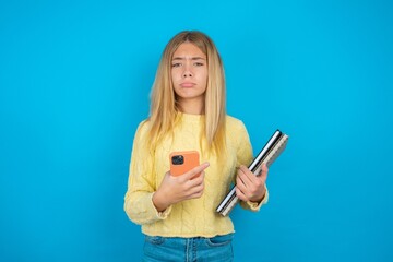 Upset dissatisfied beautiful caucasian teen girl wearing yellow sweater uses mobile software application and surfs information in internet, holds modern mobile hand