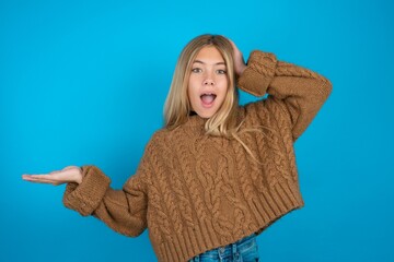 Shocked amazed surprised beautiful caucasian teen girl wearing brown sweater hold hand offering...