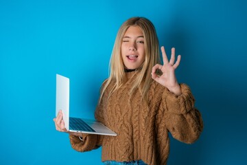 Attractive cheerful skilled beautiful caucasian teen girl wearing brown sweater using laptop showing ok-sign winkin