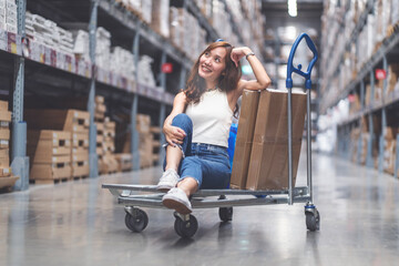 Young Asian woman in jeans sitting on a shopping cart, happy shopping for furniture in a warehouse,...