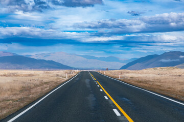 Photograph of a road heading towards a mountain range through a rural area with low level grey...