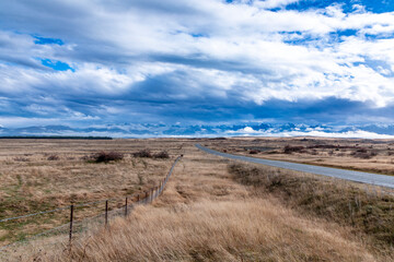 Photograph of a road heading towards a mountain range through a rural area with low level grey clouds on the South Island of New Zealand